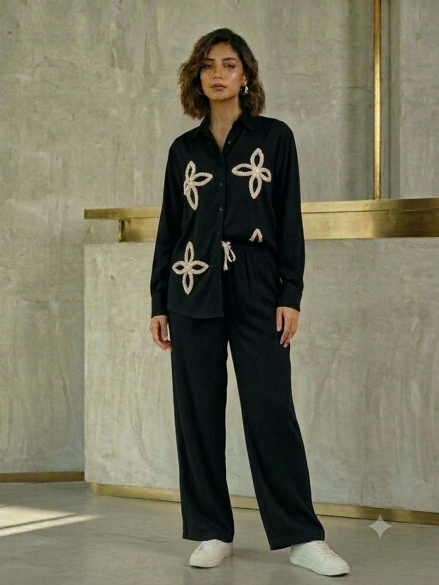 Model wearing a black long-sleeve button-front shirt with cream petal-shaped embellishments and matching wide-leg drawstring trousers
