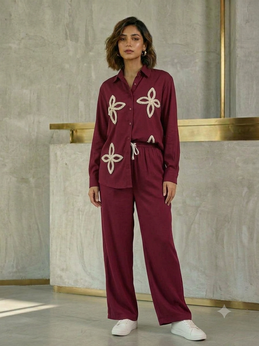 Model wearing a wine long-sleeve button-front shirt with cream petal-shaped embellishments and matching wide-leg drawstring trousers